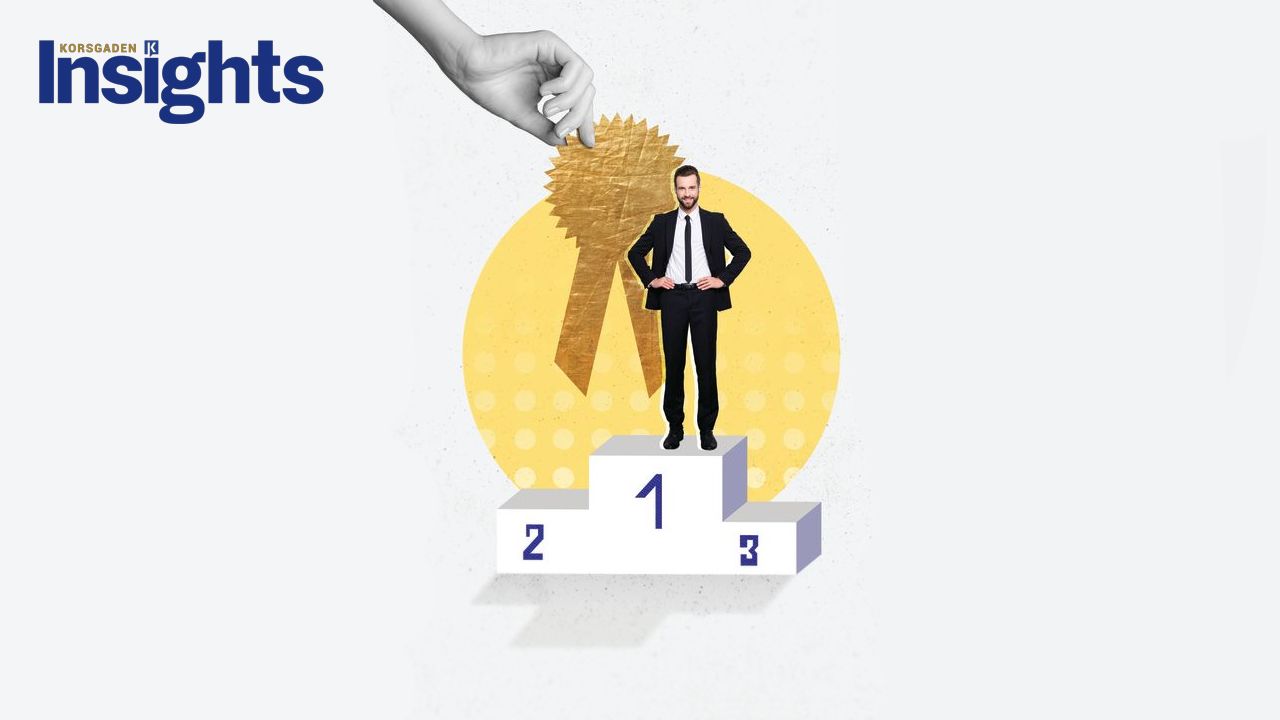 Indispensable Illustration of a confident businessman standing on a number one podium, with a large gold award ribbon being lowered behind him by a hand. The image symbolizes recognition, excellence, and being indispensable. 'Korsgaden Insights' logo appears in the top left.